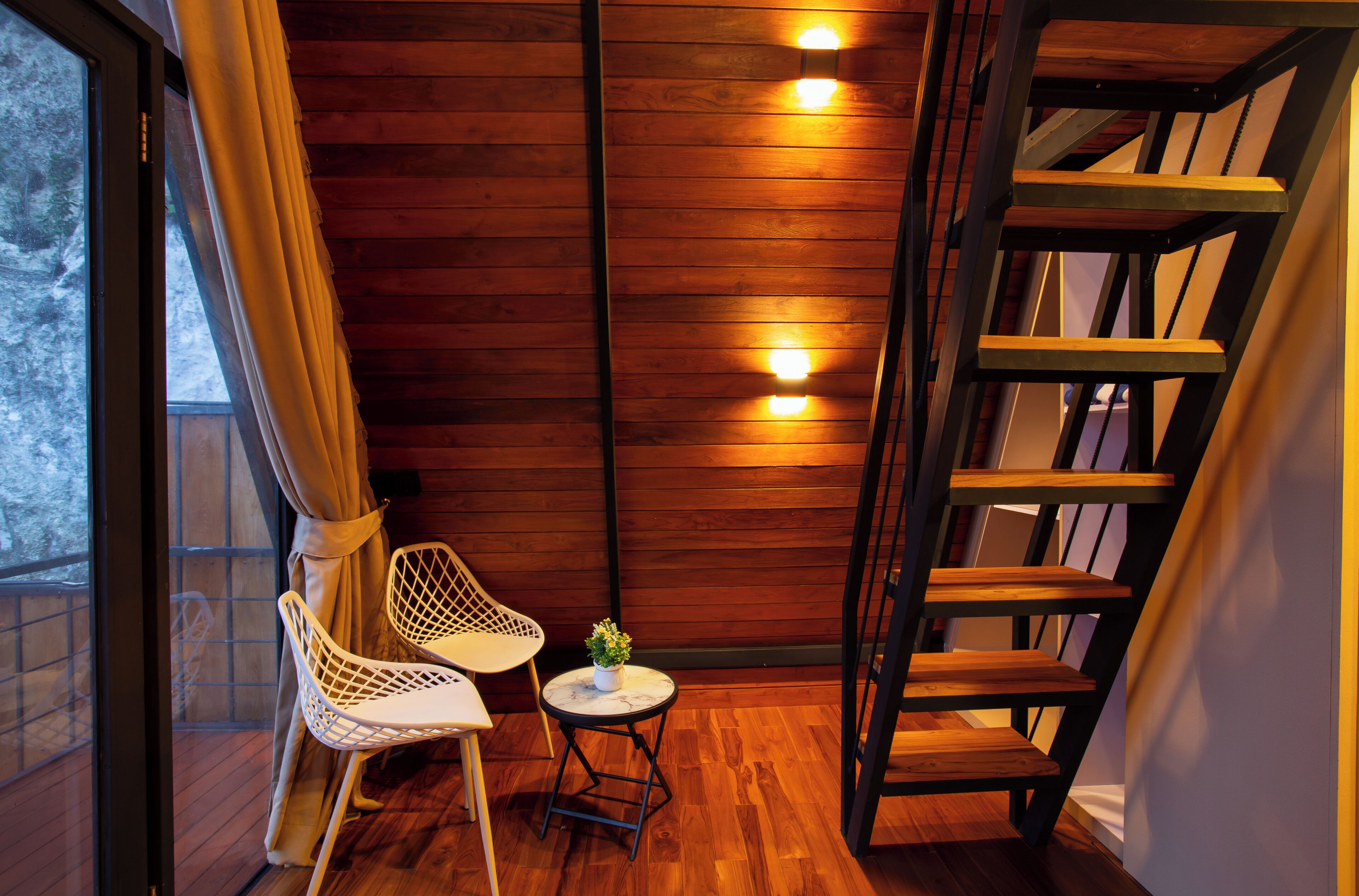 Interior de madera con una escalera, una ventana con cortinas y una pequeña mesa con una planta.
