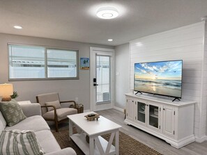 A living room with a sofa, armchair, coffee table, TV on a cabinet, and a door with blinds.