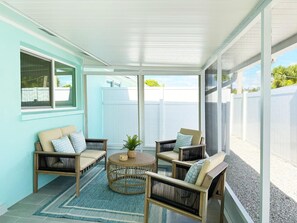A covered patio area with wicker furniture, a woven coffee table, and a view of a white fence and greenery.