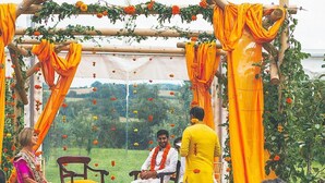 A traditional ceremony with a wooden structure, orange drapes, and a seated individual in white attire.