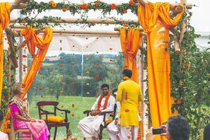 A traditional ceremony with a wooden structure, orange drapes, and a seated individual in white attire.