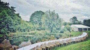 A long table set up outdoors with chairs, a tent in the background, and a pond.