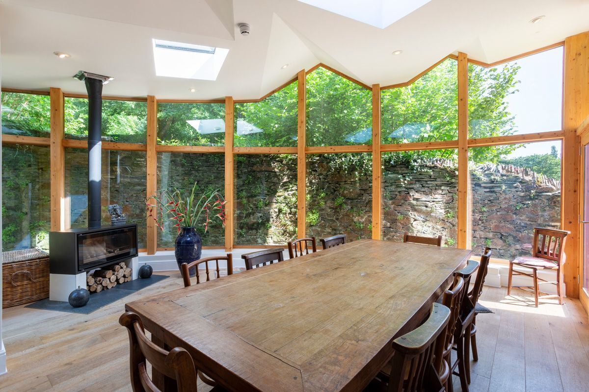 A spacious dining area with a large wooden table and chairs, a stone fireplace, and large windows offering a view of the outdoors.