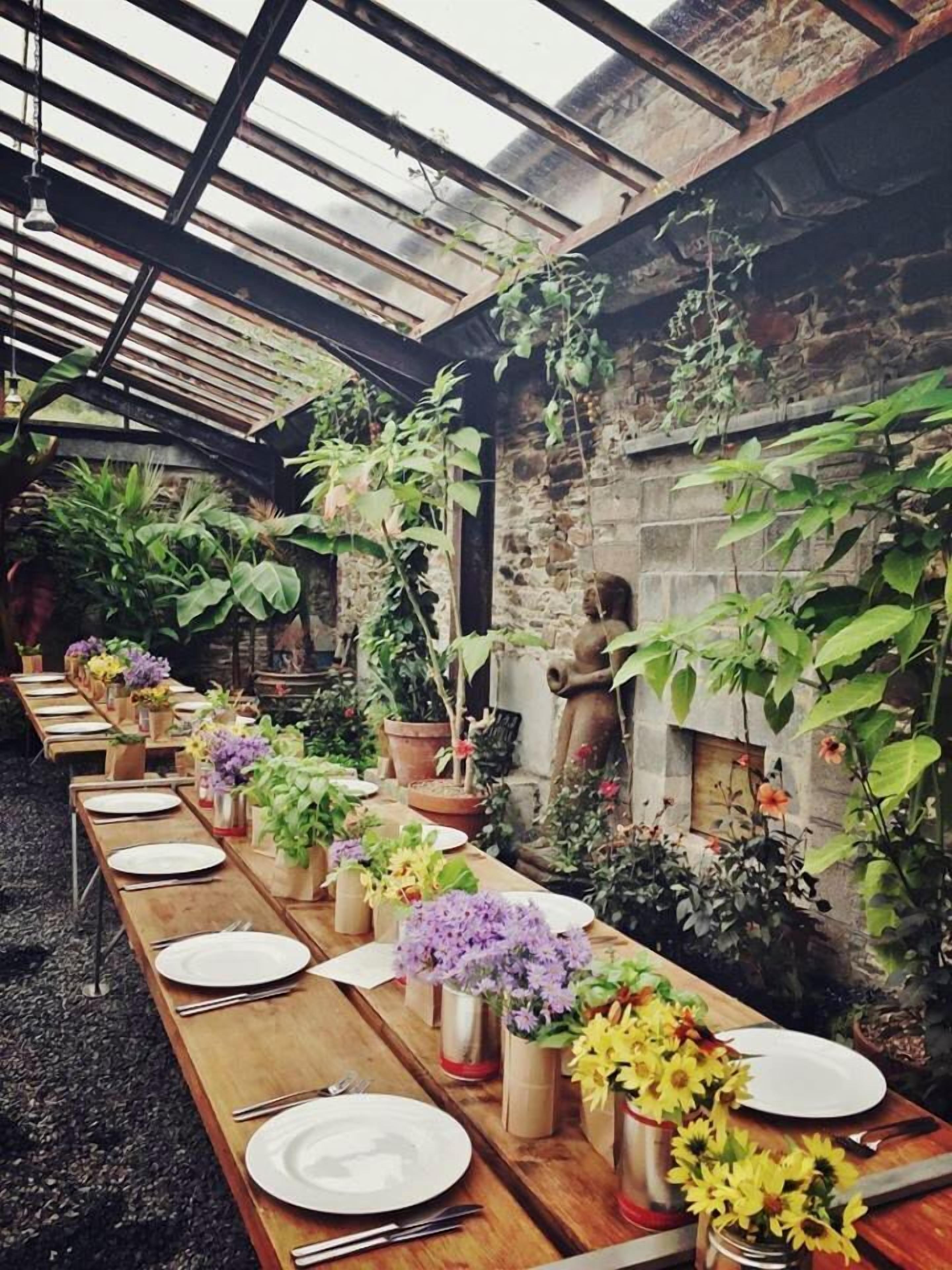 A long wooden table set for dining with potted plants and flowers, in a rustic room with stone walls and a metal roof.