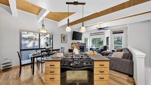 A kitchen with a central island, wooden ceiling beams, a fireplace, and a dining area with chairs and a table.