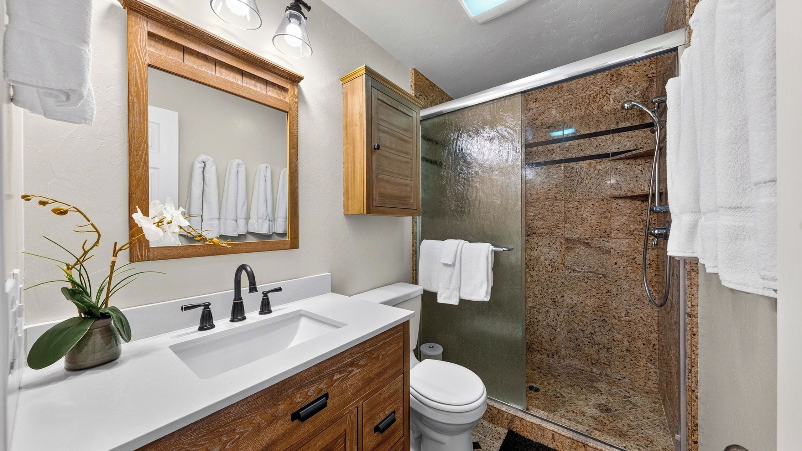 Un baño con lavamanos blanco, tocador de madera, un espejo, inodoro y cabina de ducha con mampara de vidrio.