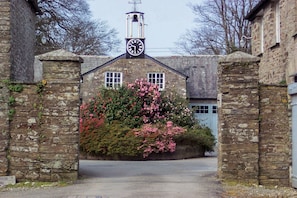Un portón de piedra con una torre de reloj, un arbusto cubierto de flores y la entrada de un edificio.
