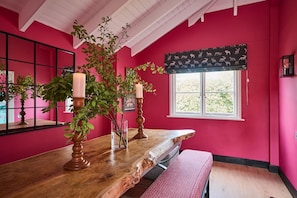 Una habitación con pared rosa, una mesa de madera con velas, un banco y una ventana con un cortinero estampado.
