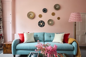 A teal sofa with red and white cushions, a wooden side table with books and pink flowers, and wall decor including circular woven items.