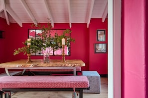 Un comedor con una mesa de madera, bancos y una pared rosa decorada con cuadros.
