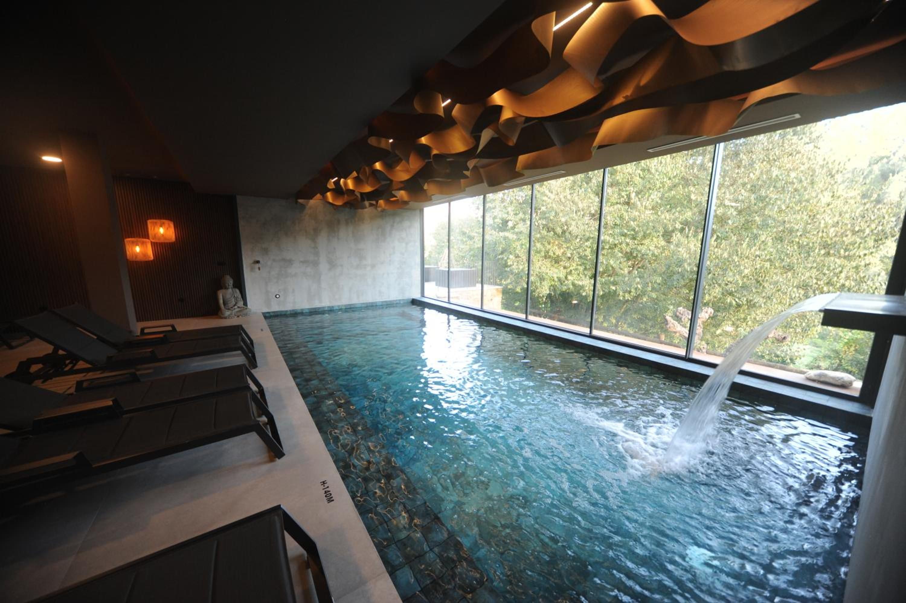 Una piscina interior con cascada, sillas de descanso y vistas a zonas verdes.