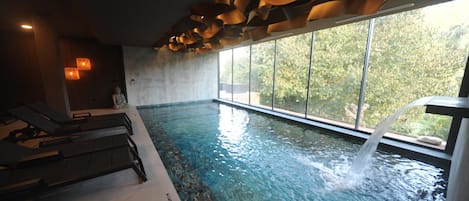 Una piscina interior con cascada, sillas de descanso y vistas a zonas verdes.