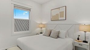 Ein Schlafzimmer mit einem Bett, zwei Nachttischen, einem Fenster mit Jalousien, einem Wandbild und einem Beistelltischchen mit Lampe.