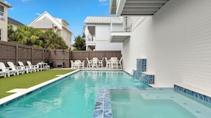Piscina com borda azulejada, rodeada por cadeiras e mesas brancas, e cerca de madeira.
