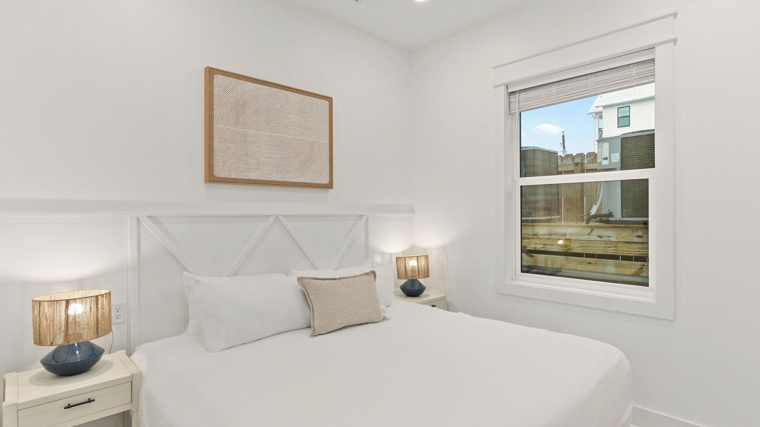 Un dormitorio con cama, dos lámparas de noche, una obra de arte en la pared y una ventana con vistas a edificios.