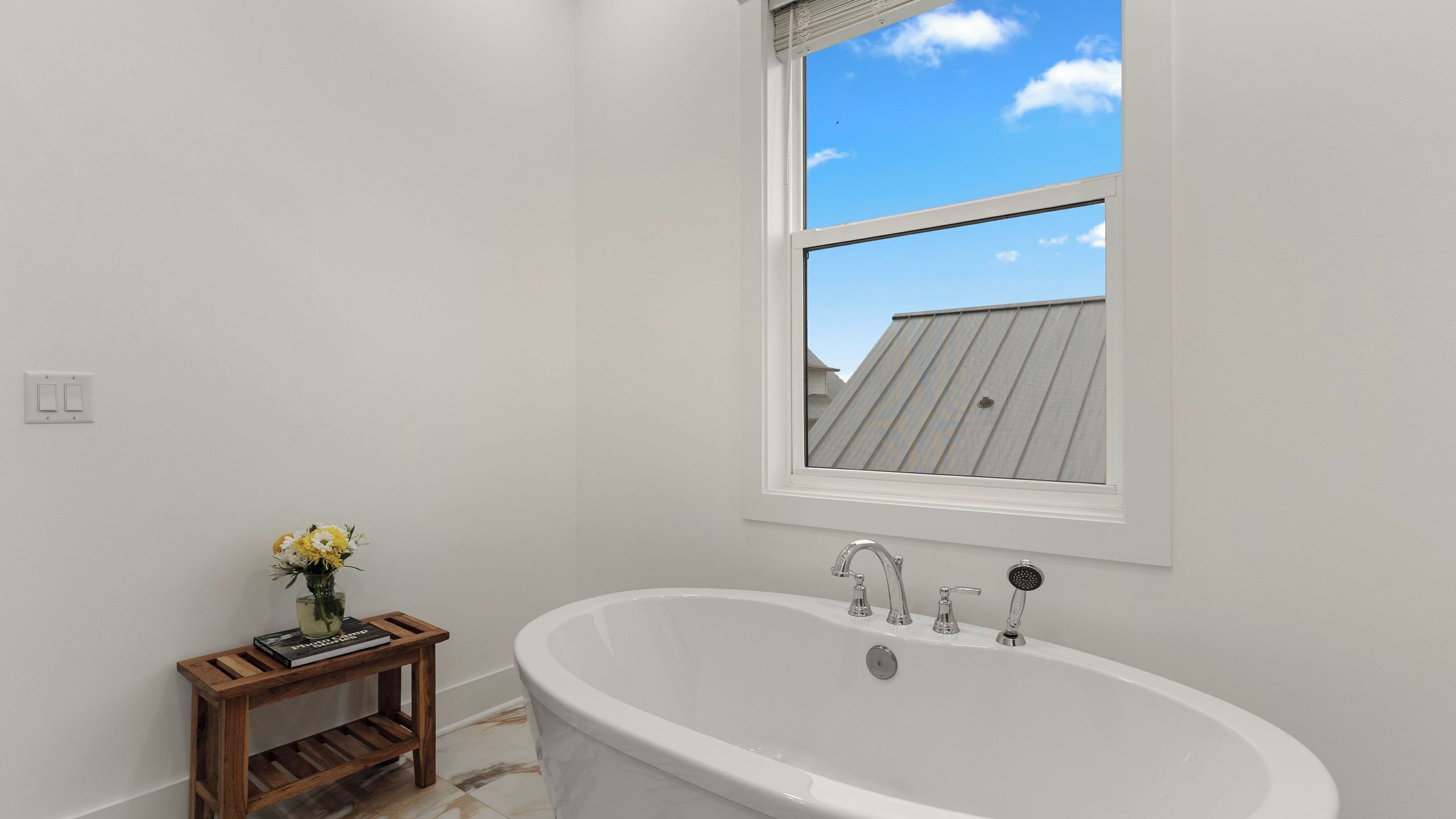A bathroom with a bathtub, a window with a view of a building, a small wooden stool with a vase of flowers, and a light switch on the wall.