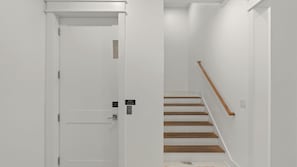 Uma porta branca com fechadura eletrônica, uma escada de madeira com corrimão e uma parede branca.