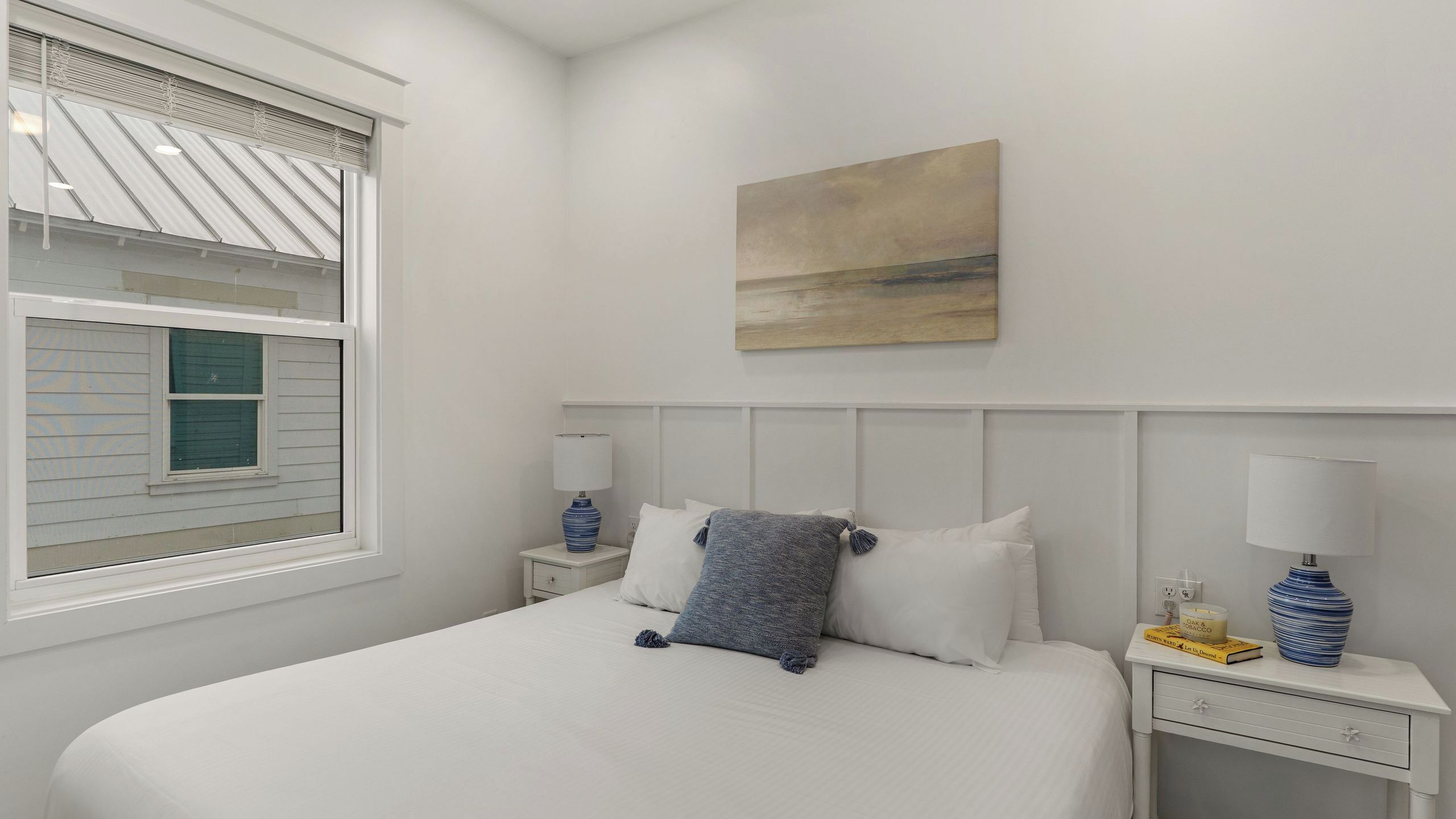 Un dormitorio con cama, dos mesitas de noche, una ventana con persianas, un cuadro en la pared y un florero sobre la mesita.