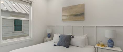 Un dormitorio con cama, dos mesitas de noche, una ventana con persianas, un cuadro en la pared y un florero sobre la mesita.