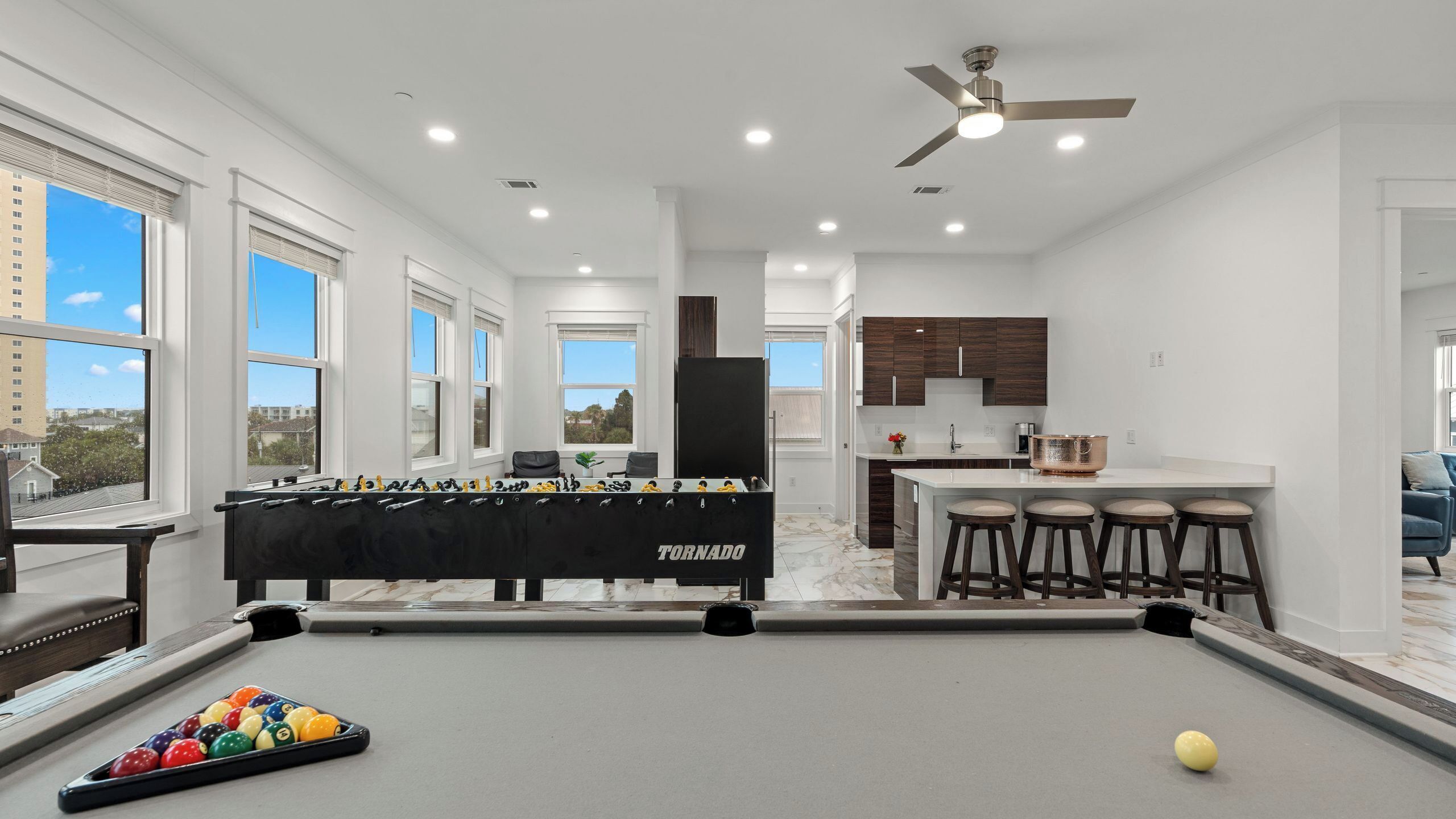 Une cuisine moderne avec des meubles en bois foncé, un îlot central doté de tabourets et une table de billard, avec une table de baby-foot en premier plan.