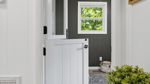 Un couloir avec une porte blanche, une fenêtre donnant sur des arbres, un mur blanc et une plante posée sur une table.