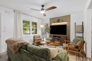 Ein Wohnzimmer mit einer Couch, Sesseln, einem Couchtisch, einem Fernseher, einem Kamin und einem Deckenventilator.