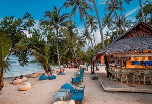 Eine Strandbar mit Außenbereich, Palmen und einer Hütte mit Reetdach.