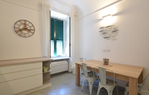 A room with a wooden table, white chairs, a wall clock, a window with blinds, and a wall-mounted light fixture. - 2 bedroom beautiful apartment in Loano (Loano)