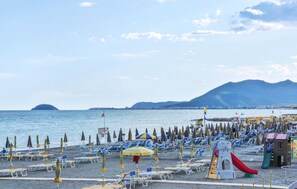 Ein Strand mit Liegestühlen und Sonnenschirmen, einem Spielplatz und einem Berg im Hintergrund.