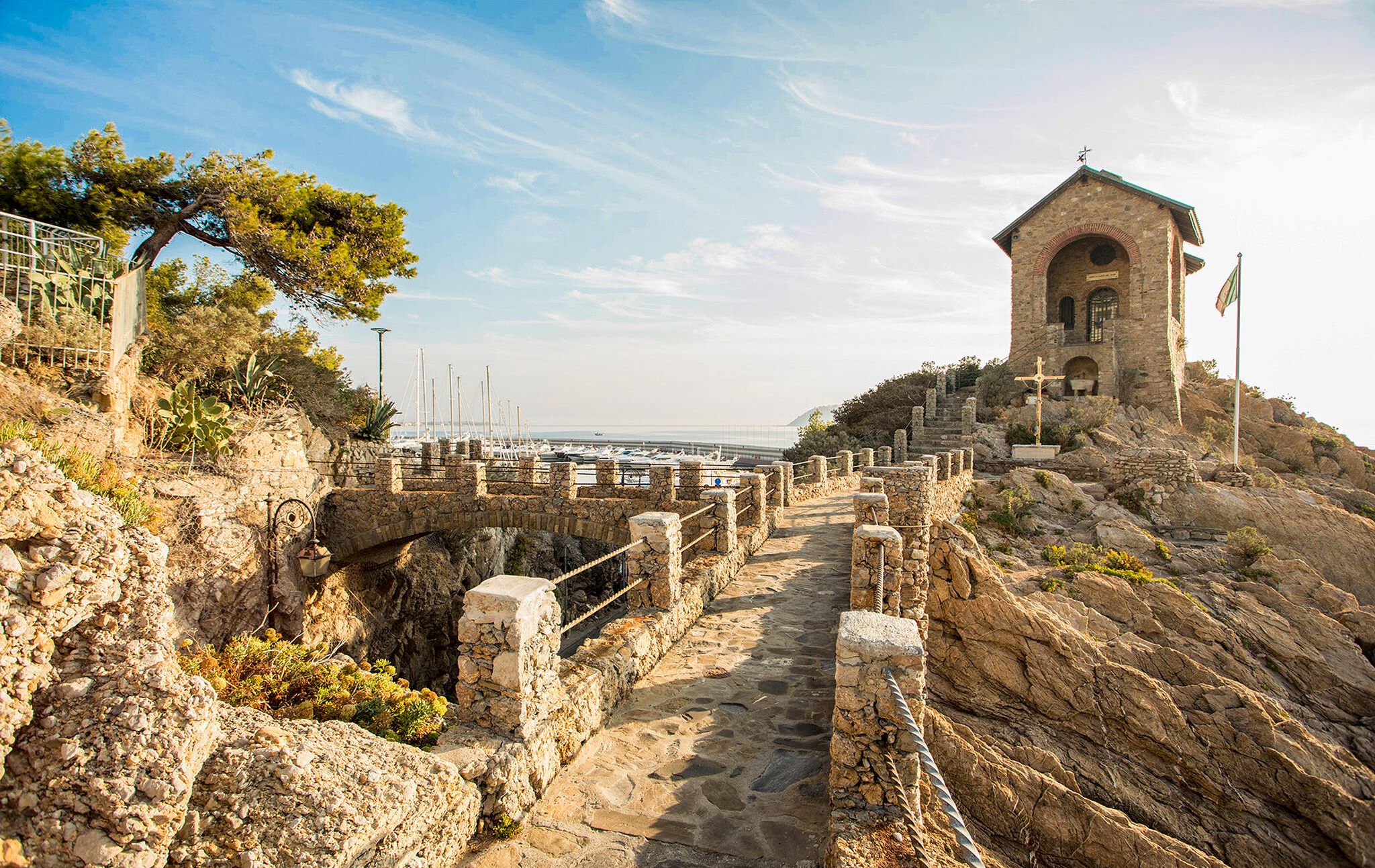 A stone pathway leading to a historical building with a bell tower, overlooking a marina with boats.