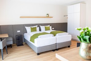 Una habitación de hotel moderna con una cama tapizada en gris, ropa de cama blanca, una mesita de noche pequeña, una silla, una lámpara de noche, un armario y una planta en maceta.