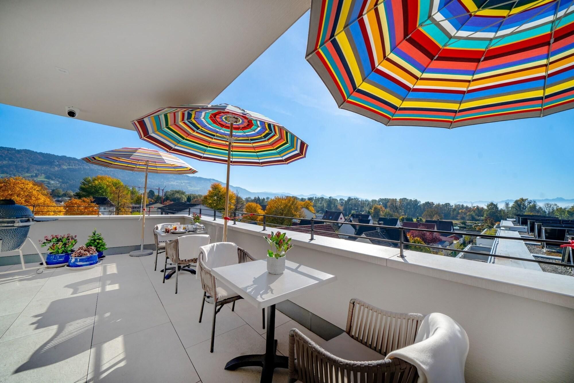 Ein Balkon mit Sitzgelegenheiten im Freien, großen, gestreiften Sonnenschirmen und Blick auf eine gebirgige Landschaft.