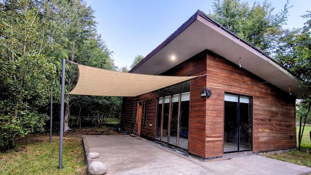 Una cabaña moderna de madera con un patio de concreto, ventanas amplias y un toldo.