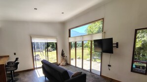 Un salón amplio con un sofá negro, un televisor montado en la pared y ventanas grandes con vistas al exterior.