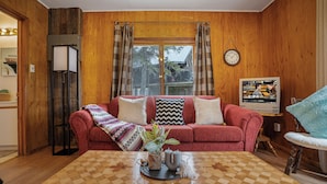 A cozy living room with a wooden interior, a red sofa with patterned cushions, a coffee table with a chessboard, and a television displaying a program.