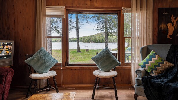 A room with wooden walls, a large window with curtains, two rocking chairs with cushions, a television, and a chair with patterned cushions.