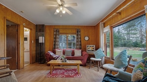 A wooden-paneled room with a ceiling fan, a red sofa, a wooden coffee table, and a view of a forest through large windows.