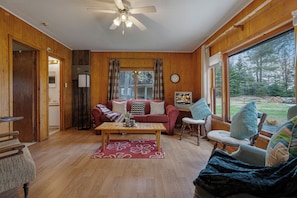 Une pièce aux murs lambrissés, dotée d’un ventilateur de plafond, d’un canapé rouge, d’une table basse en bois et offrant une vue sur une forêt grâce à de grandes fenêtres.