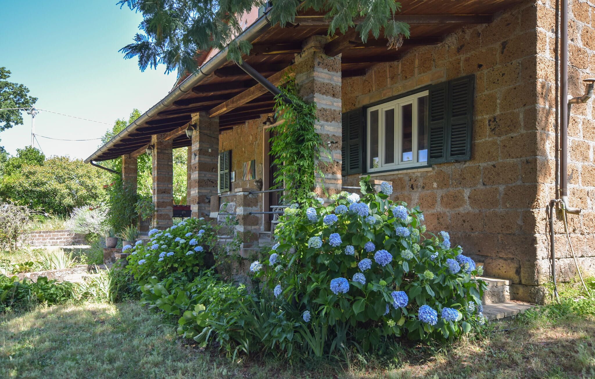 Un edificio de piedra rústica con un porche cubierto, una ventana con contraventanas y arbustos de hortensias en primer plano.