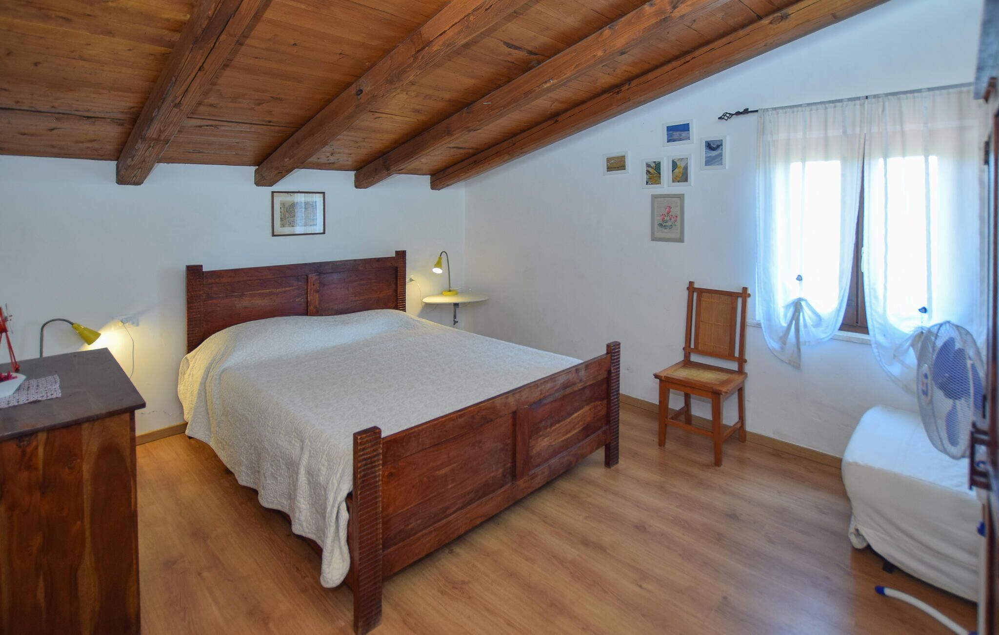 A bedroom with a wooden bed, bedside table, lamp, chair, and a bedside fan.