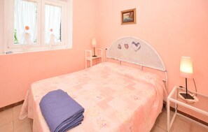 Ein Schlafzimmer mit einem Bett, einem Nachttisch, einer Lampe, einem Fenster mit Vorhängen und einem Bild an der Wand.