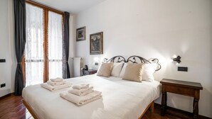 A bedroom with a bed, two wall-mounted lights, a radiator, and a window with curtains. - Italianway Easy - Corso San Gottardo 5 (Milano)