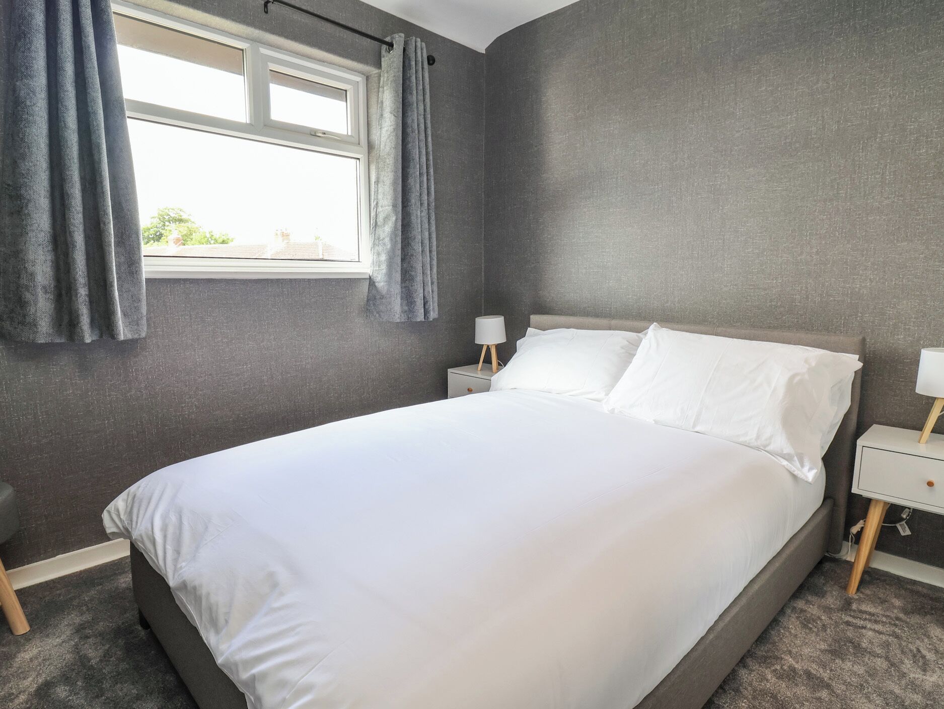 Un dormitorio con una cama grande, paredes grises, una ventana con cortinas y mesitas de noche con lámparas.