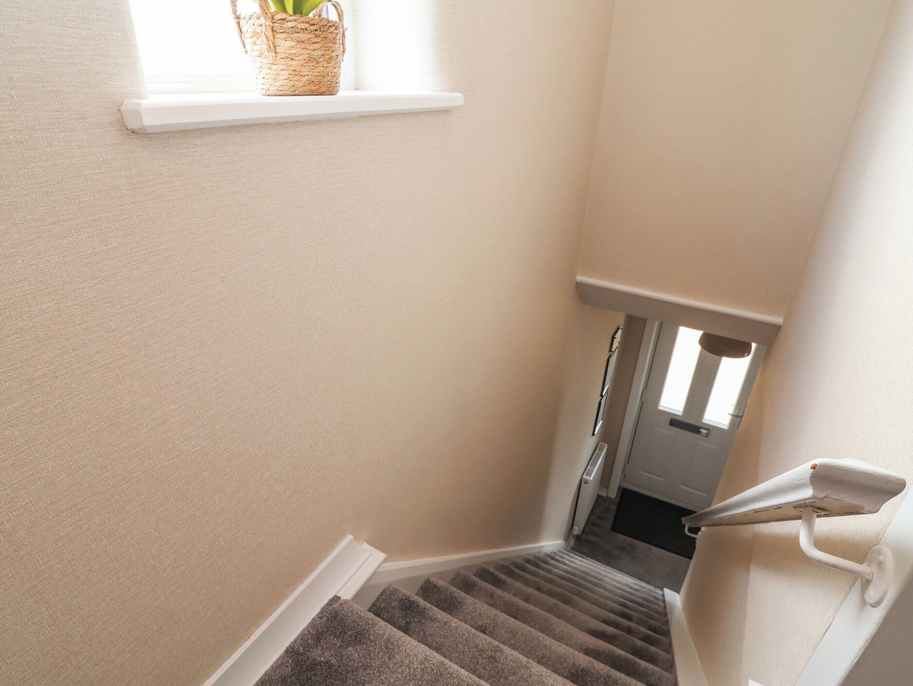 Una escalera alfombrada con barandilla blanca, una ventana pequeña con una cesta, y una puerta en la parte superior.