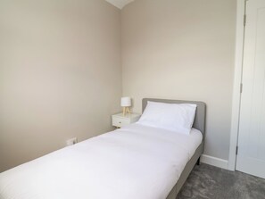 Una cama individual con cabecera gris, una mesita de noche blanca con lámpara y suelo cubierto de alfombra gris.