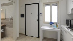 Een hotelkamer met een badkamer, een wit bureau met een vaas, een magnetron en een raam met jaloezieën.