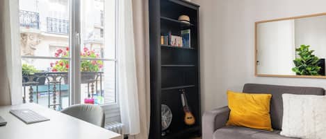 Een woonkamer met een grijze bank, een zwarte boekenkast, een raam met uitzicht op gebouwen, en een radiator.