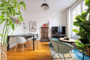 Ein modernes Wohnzimmer mit Holzboden, einem Esstisch mit Stühlen, einem Kamin, einem Fernseher und großen Grünpflanzen.