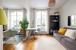 Ein Wohnzimmer mit einer Couch, einem Esstisch, Stühlen, einer Topfpflanze, einem Bücherregal und einem Fenster mit Blick auf Gebäude.