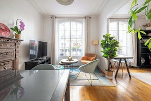 Ein Wohnzimmer mit Fernseher, einer Couch, einem Esstisch und großen Fenstern.